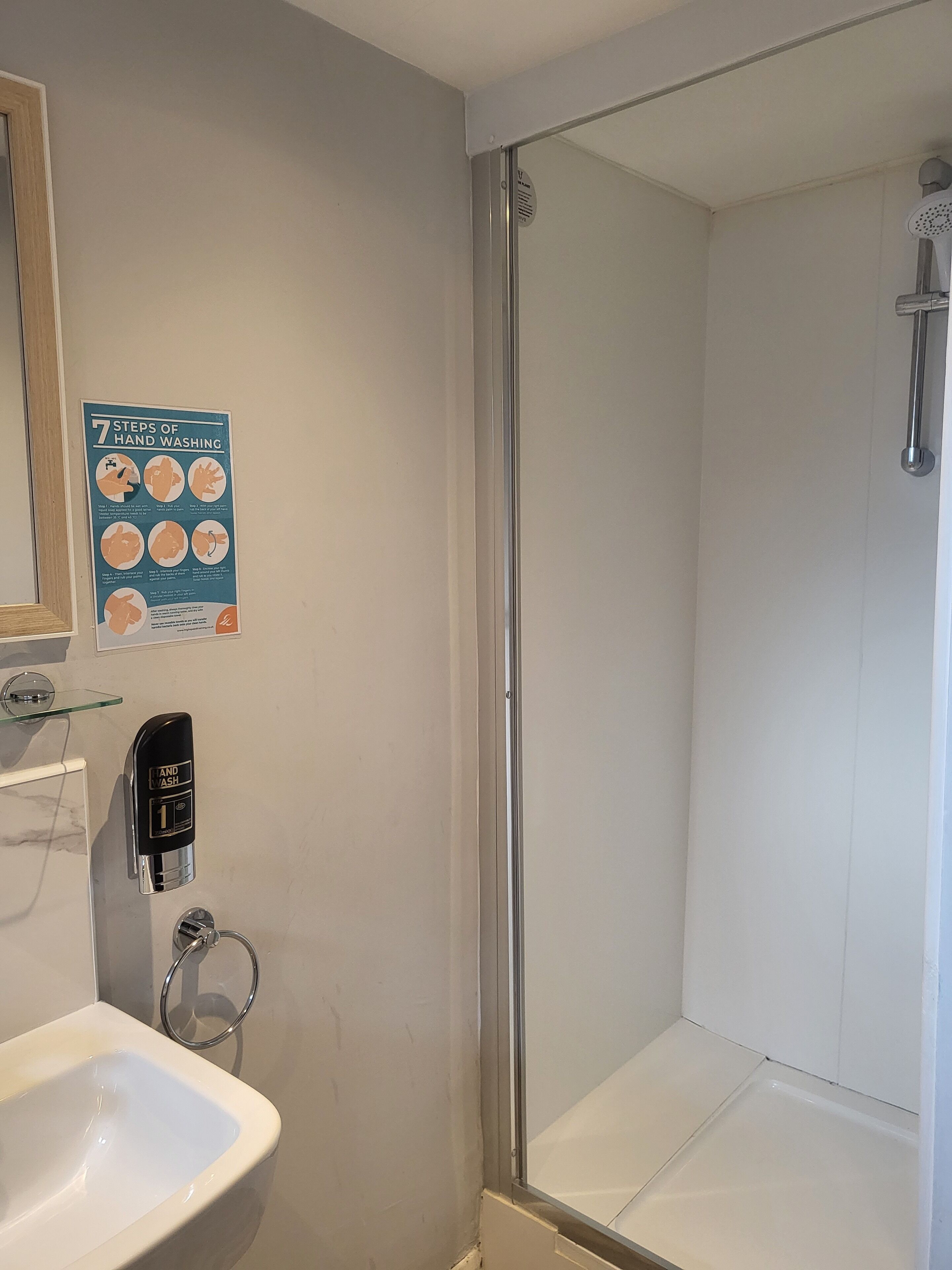 A bathroom with a glass shower enclosure, a sink, a soap dispenser, and a poster illustrating hand washing steps.
