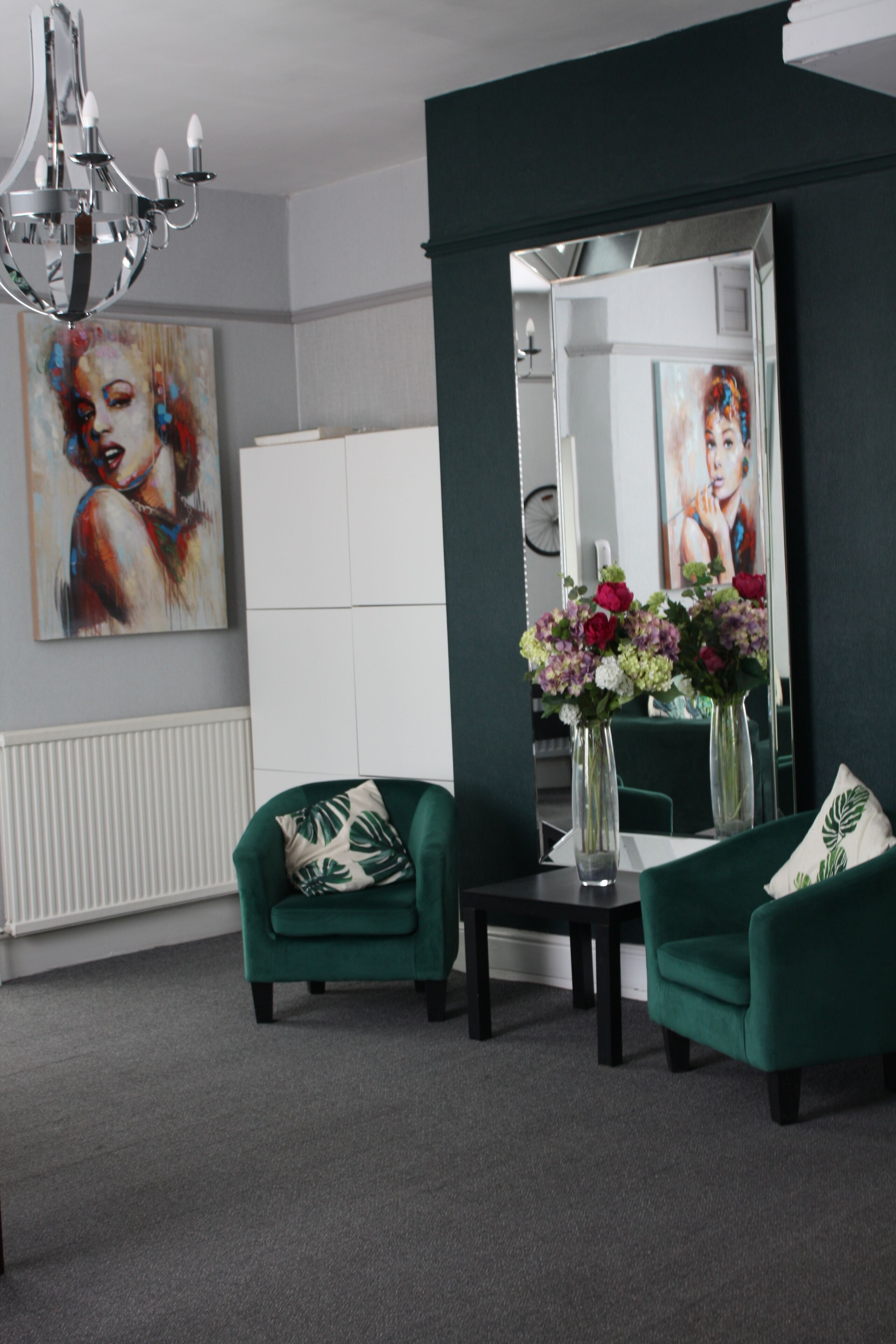 A modern living room with two green armchairs, a small table with a vase of flowers, a chandelier, and a large mirror reflecting the room.