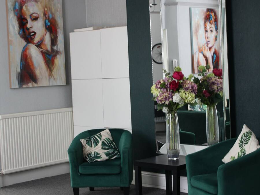 A modern living room with two green armchairs, a small table with a vase of flowers, a chandelier, and a large mirror reflecting the room.