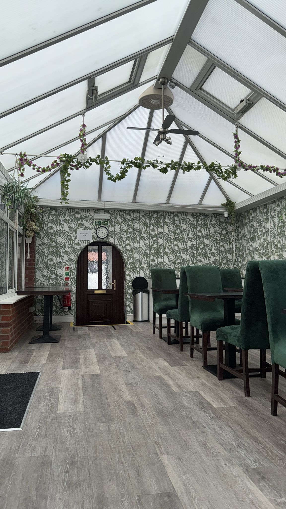 A greenhouse with a wooden floor, green chairs, a clock, and a door with a sign.