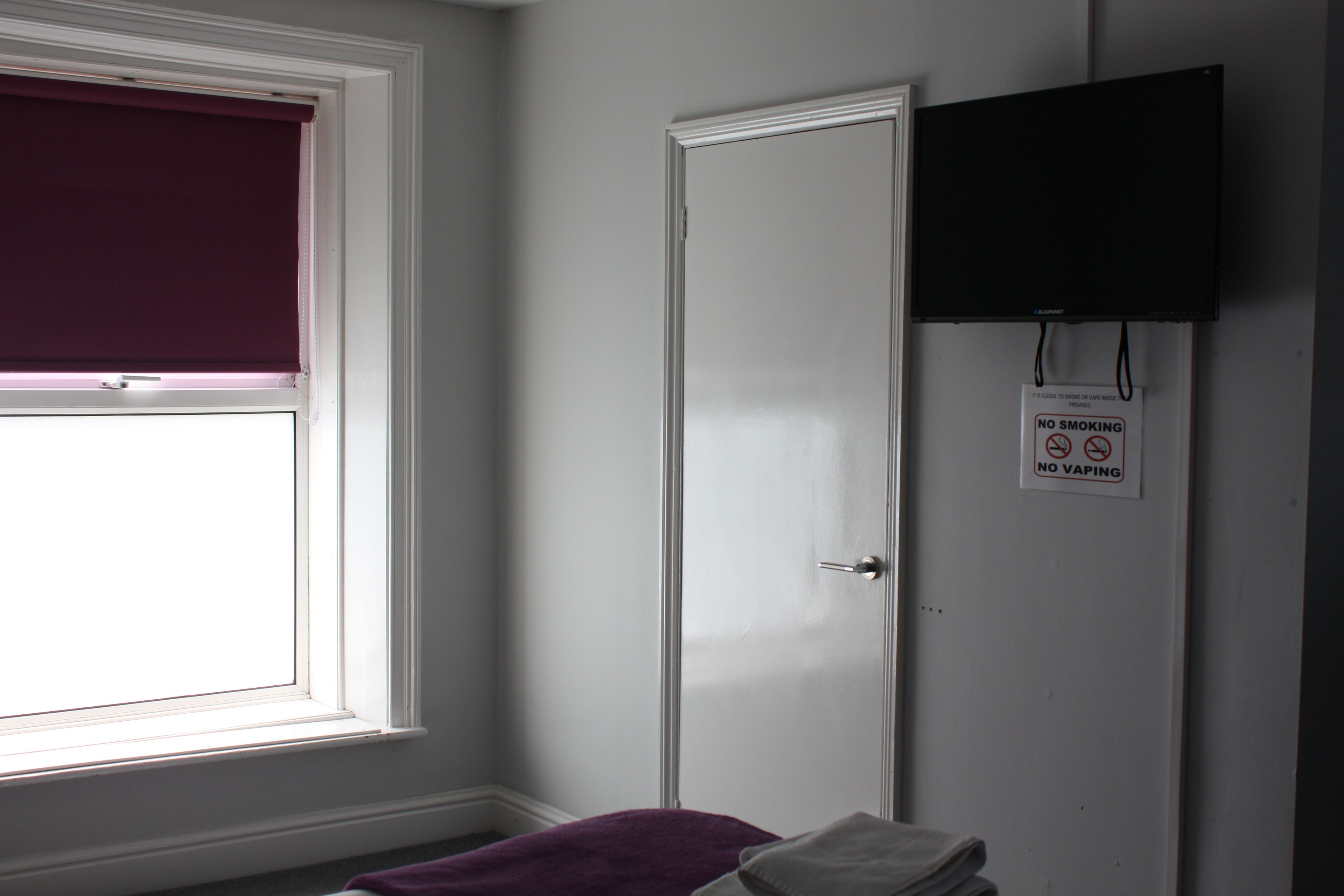 A room with a closed door, a window with a purple blind, and a no smoking/vaping sign on the wall.