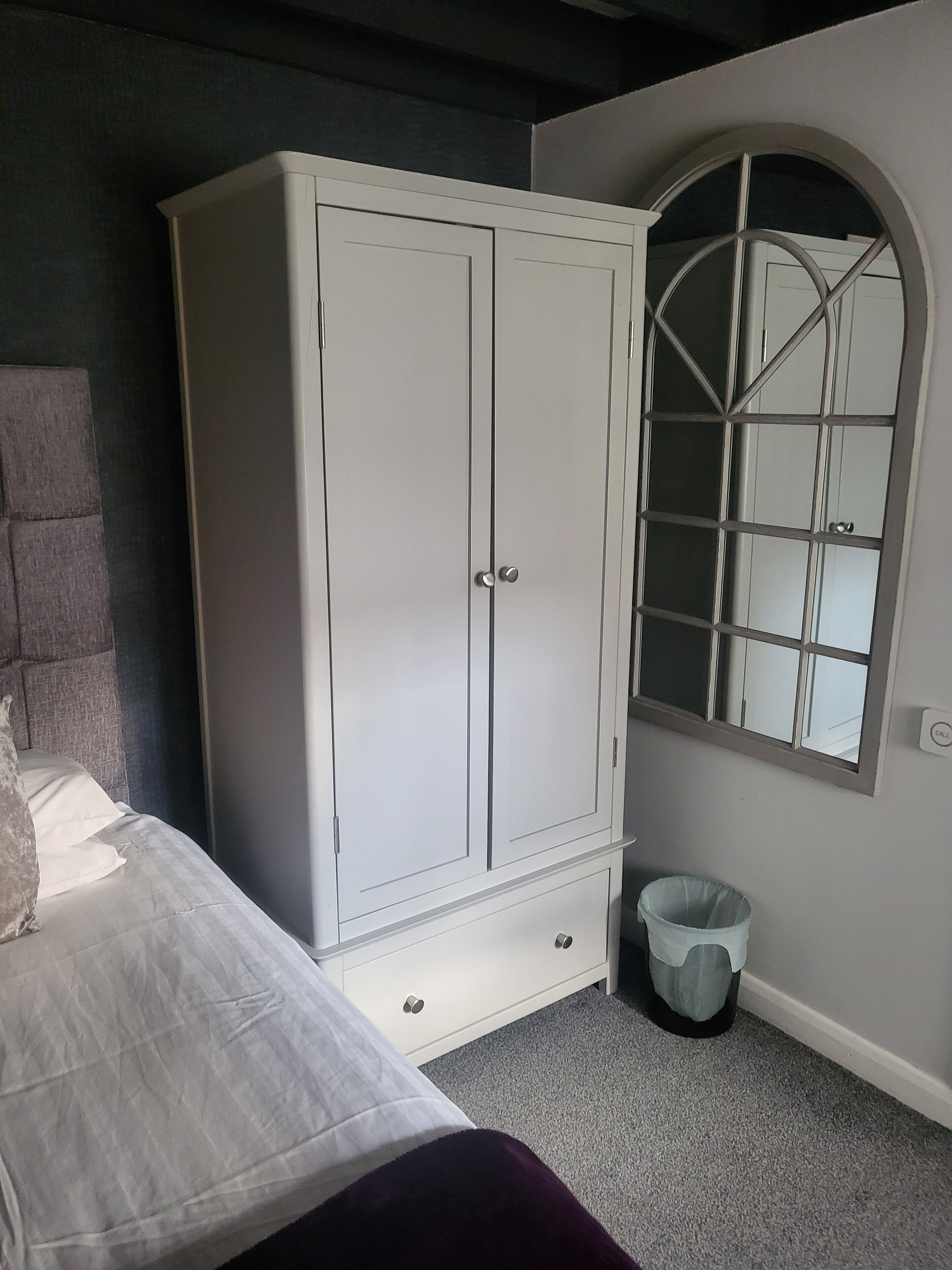 A bedroom with a bed, a white wardrobe, a small trash bin, and a mirror with an arched top.