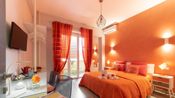 A modern hotel room with a glass desk, a bed with orange bedding, and a balcony with red curtains. - B&B La Maison de Jo (Rome)
