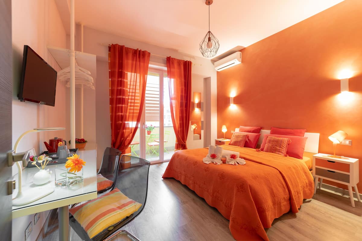 Quarto de hotel moderno com escrivaninha de vidro, cama com roupa de cama laranja e varanda com cortinas vermelhas.