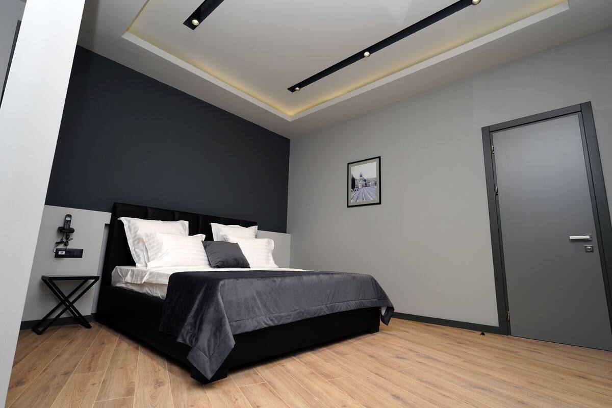 Ein modernes Schlafzimmer mit einem schwarzen Bett, weißen Kissen, einem schwarzen Nachttisch und einem gerahmten Bild an der Wand.