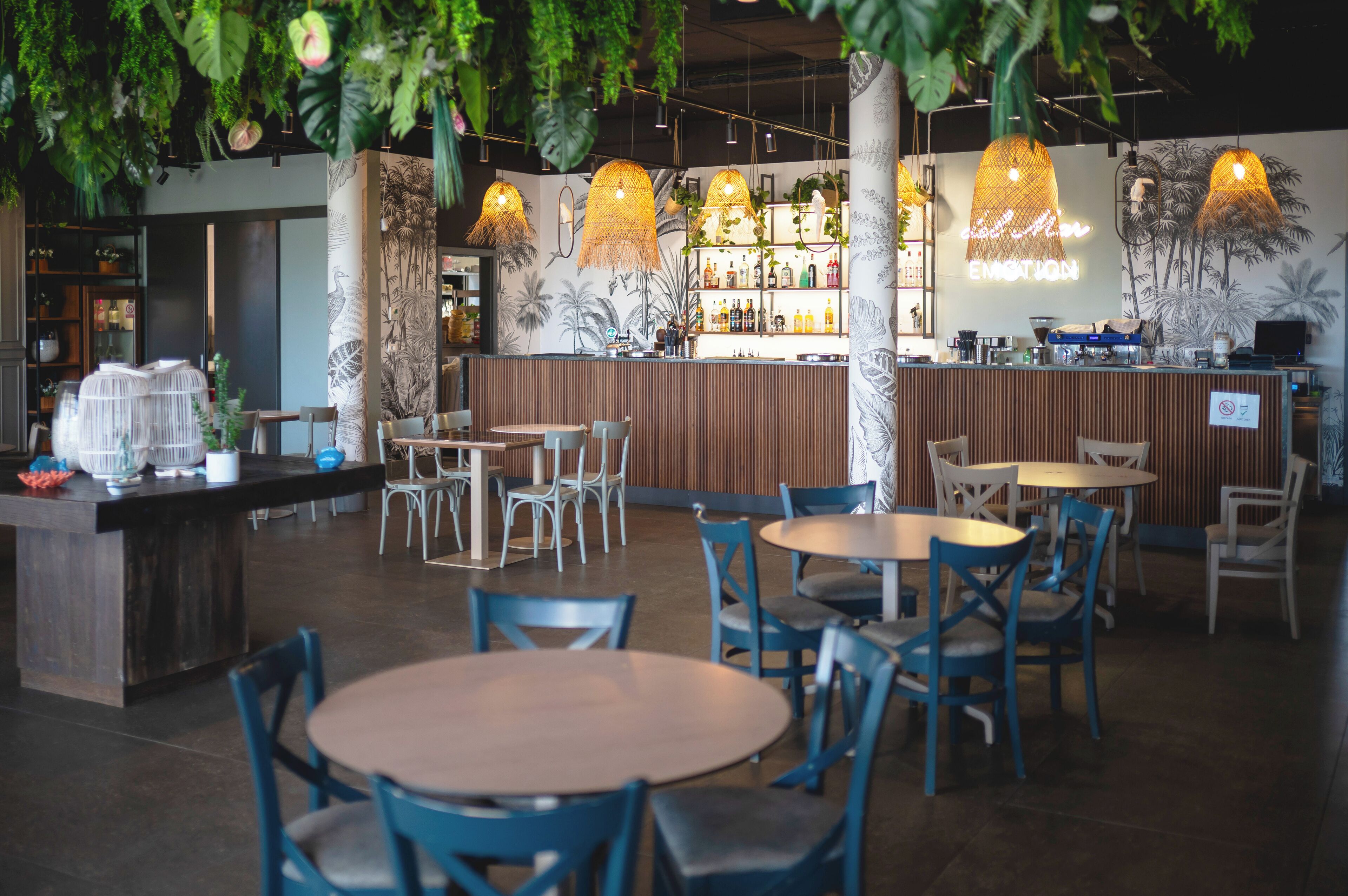 L’intérieur d’un restaurant présente des tables et des chaises rondes, un bar avec des étagères remplies de bouteilles et des plantes suspendues.