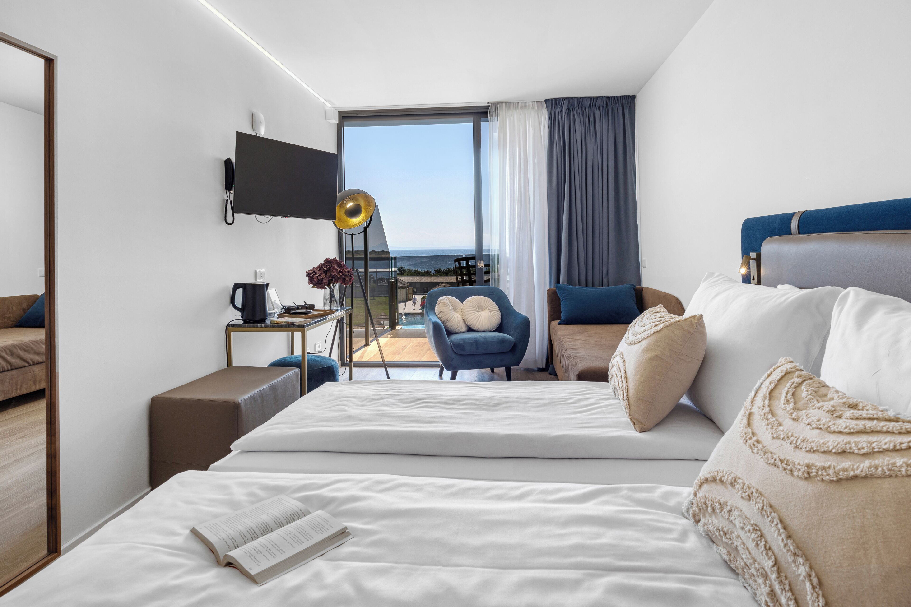 A modern hotel room with a large bed, a desk, a chair, a TV, and a balcony with a view.