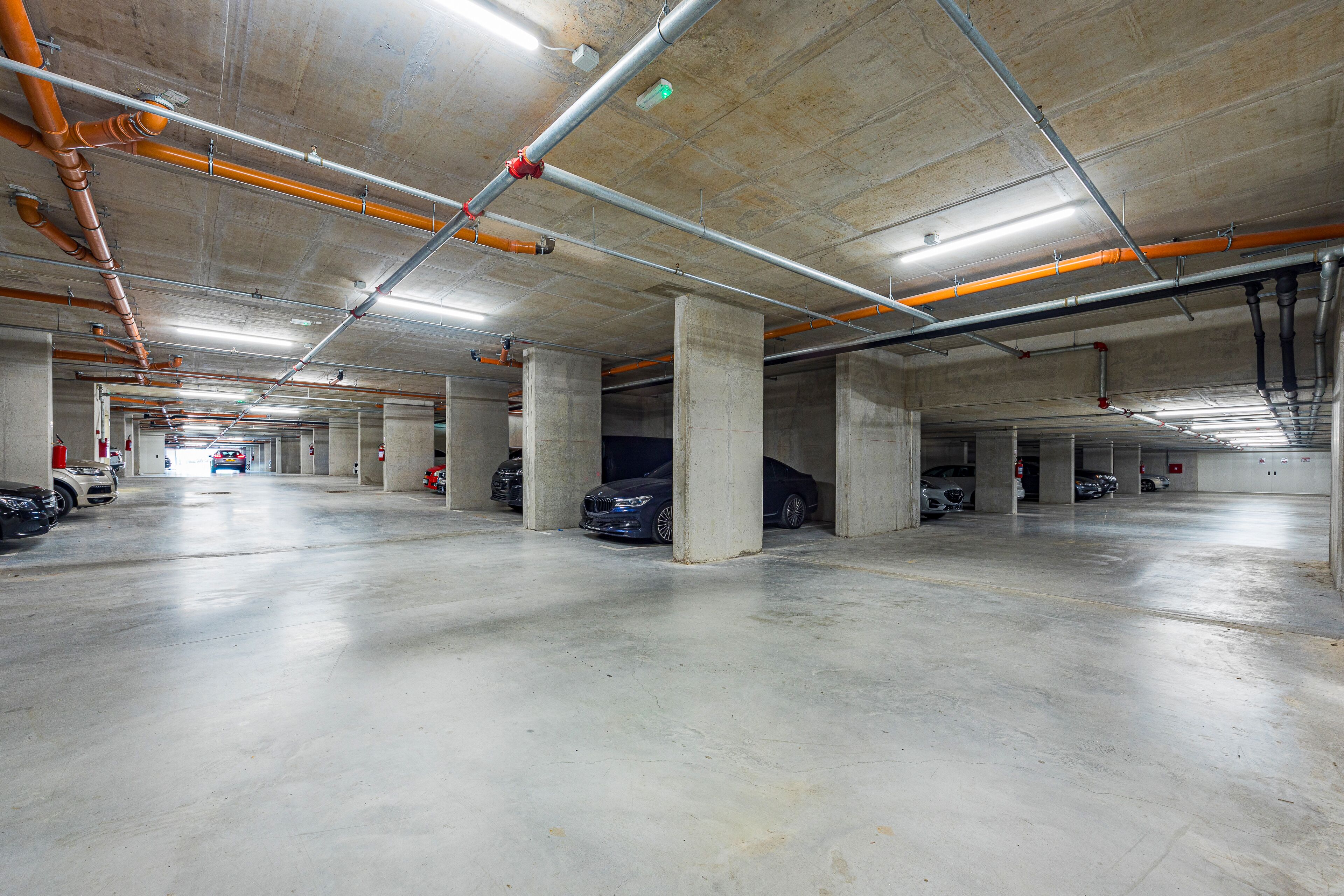 A multi-level underground parking garage with concrete pillars, overhead pipes, and several parked cars.