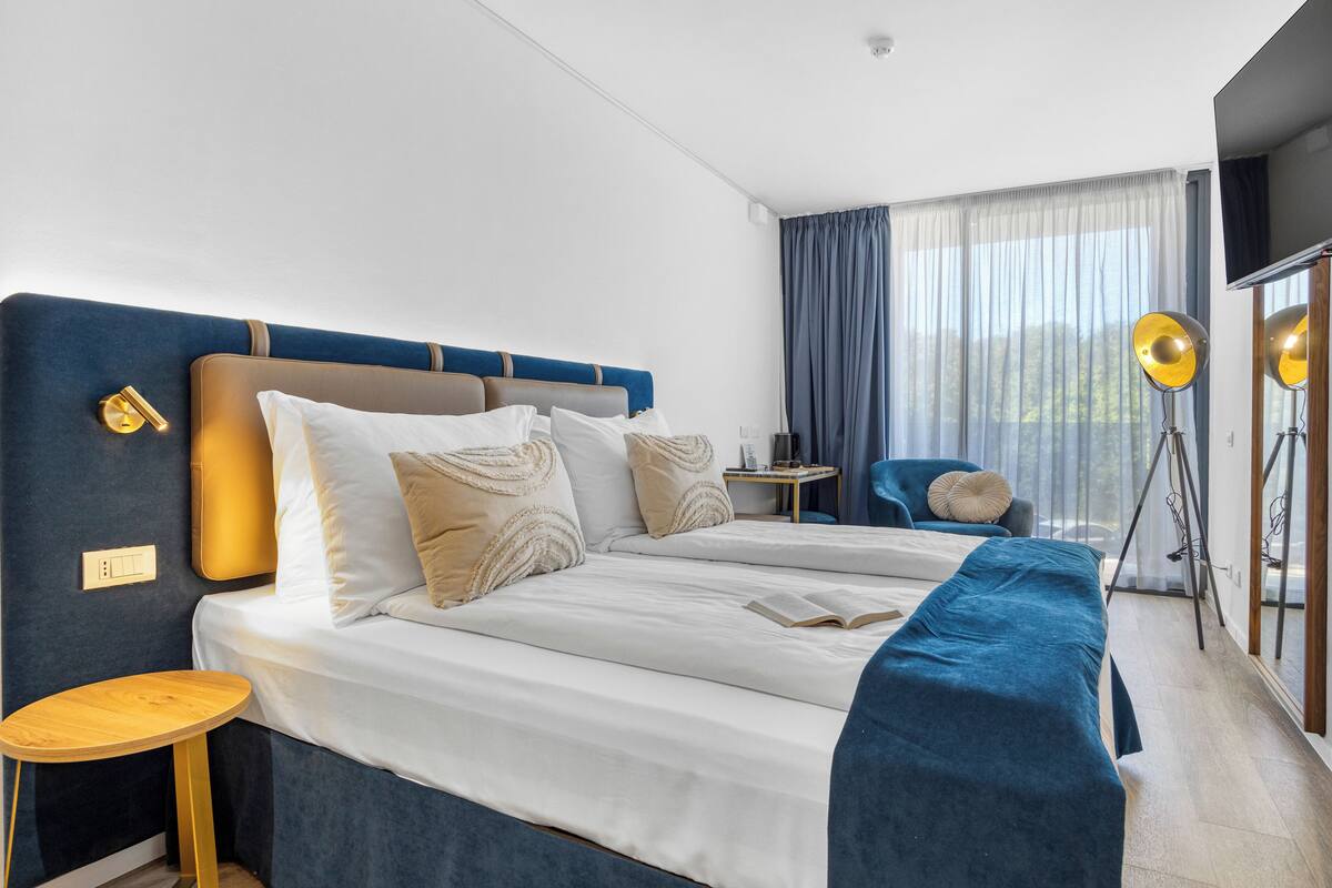 A hotel room with a large bed, bedside tables, a chair, a TV, and a window with curtains.