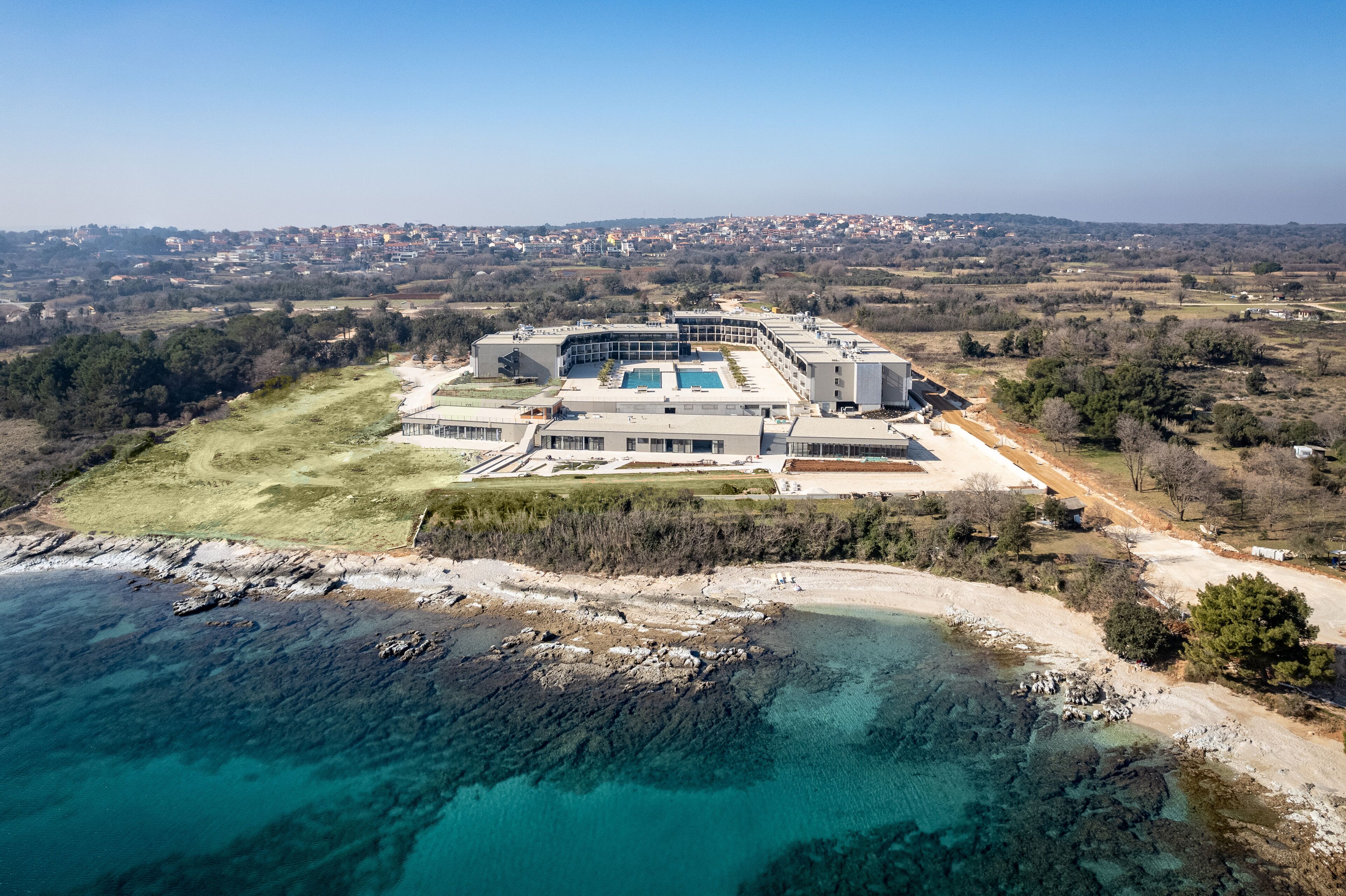 An aerial view of a coastal building complex with a swimming pool, surrounded by a mix of natural and developed land.
