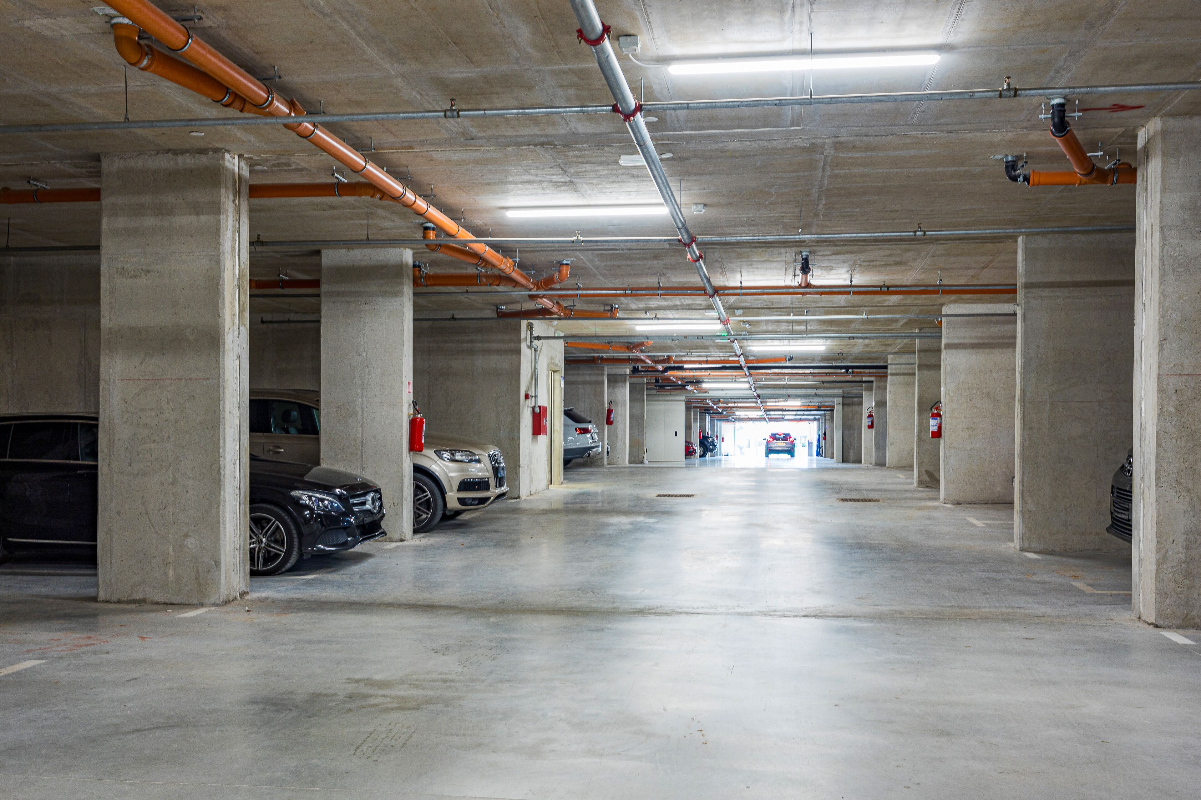 Un parking à plusieurs niveaux, avec des piliers en béton, des tuyaux apparents et des extincteurs.