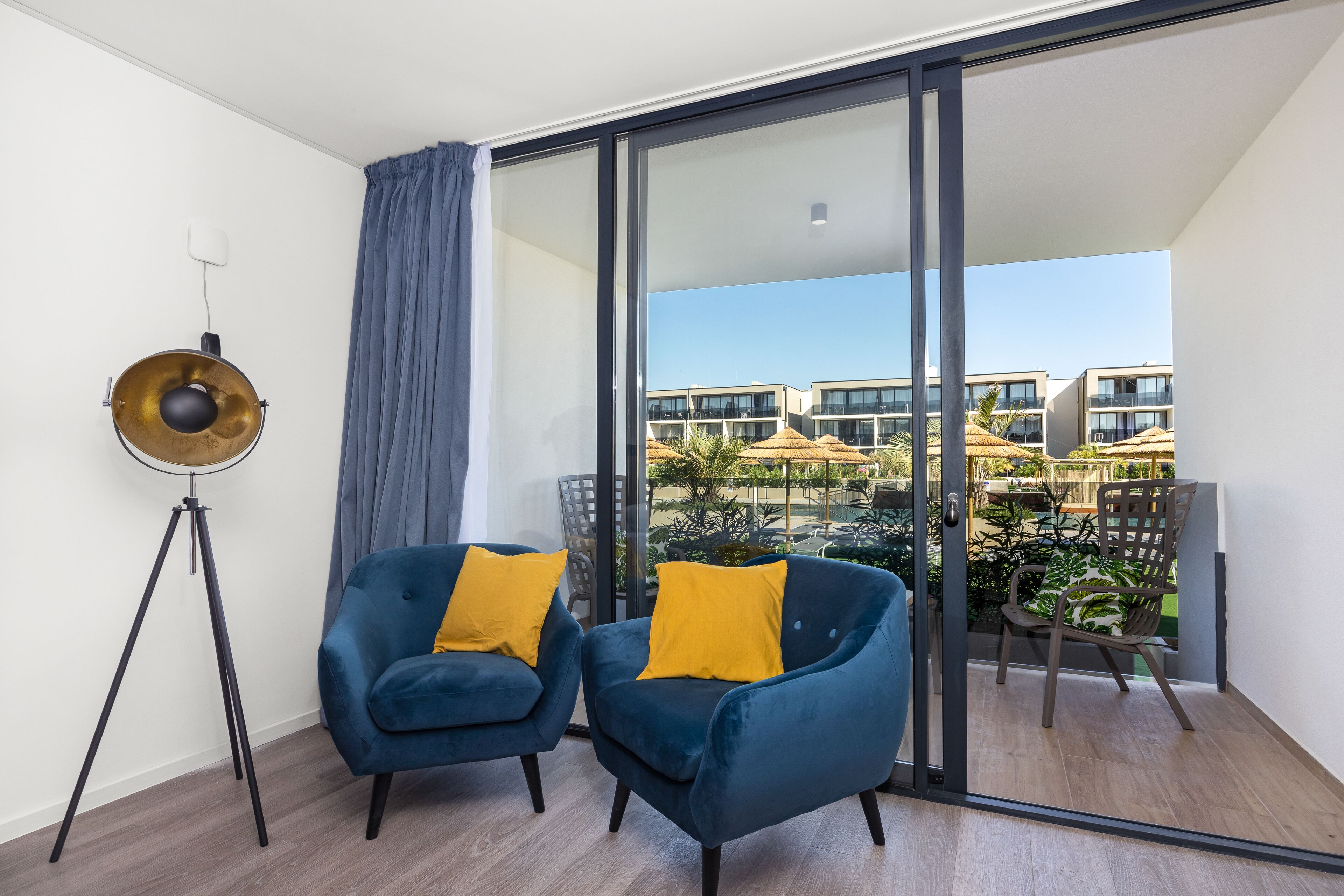A balcony with two blue armchairs and a floor lamp, overlooking a pool area with umbrellas.