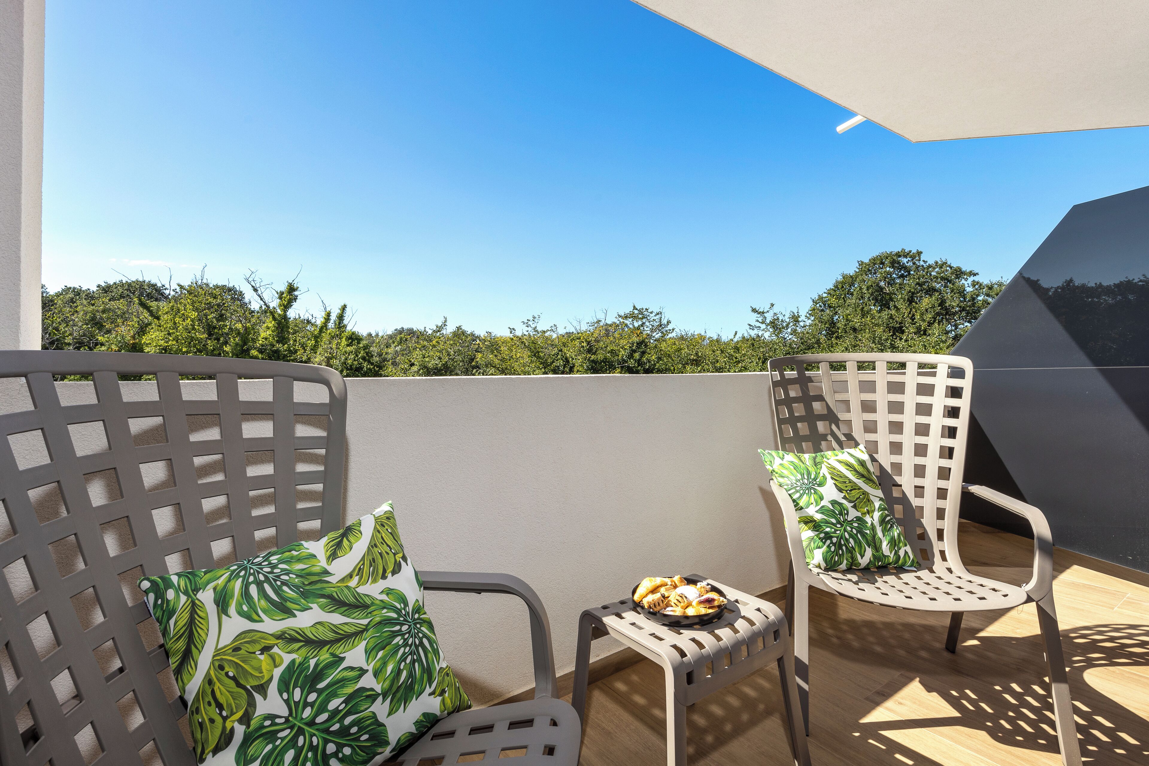 A balcony with two chairs, one with a leaf-patterned cushion and a small table with a plate of food.