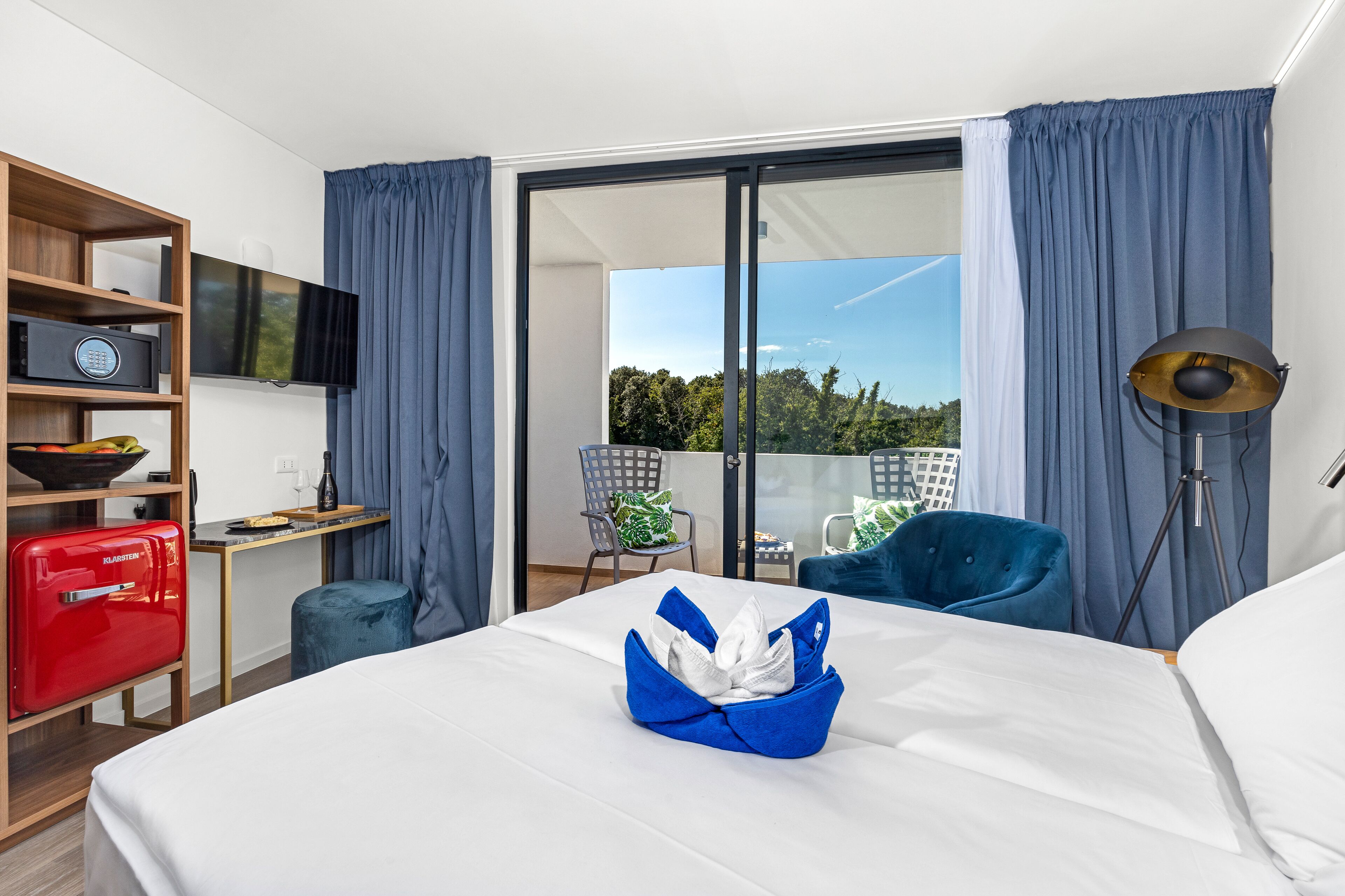 Hotelkamer met een bed, een televisie, een bureau, een rode koffer, een blauwe stoel en een balkon met uitzicht op bomen.