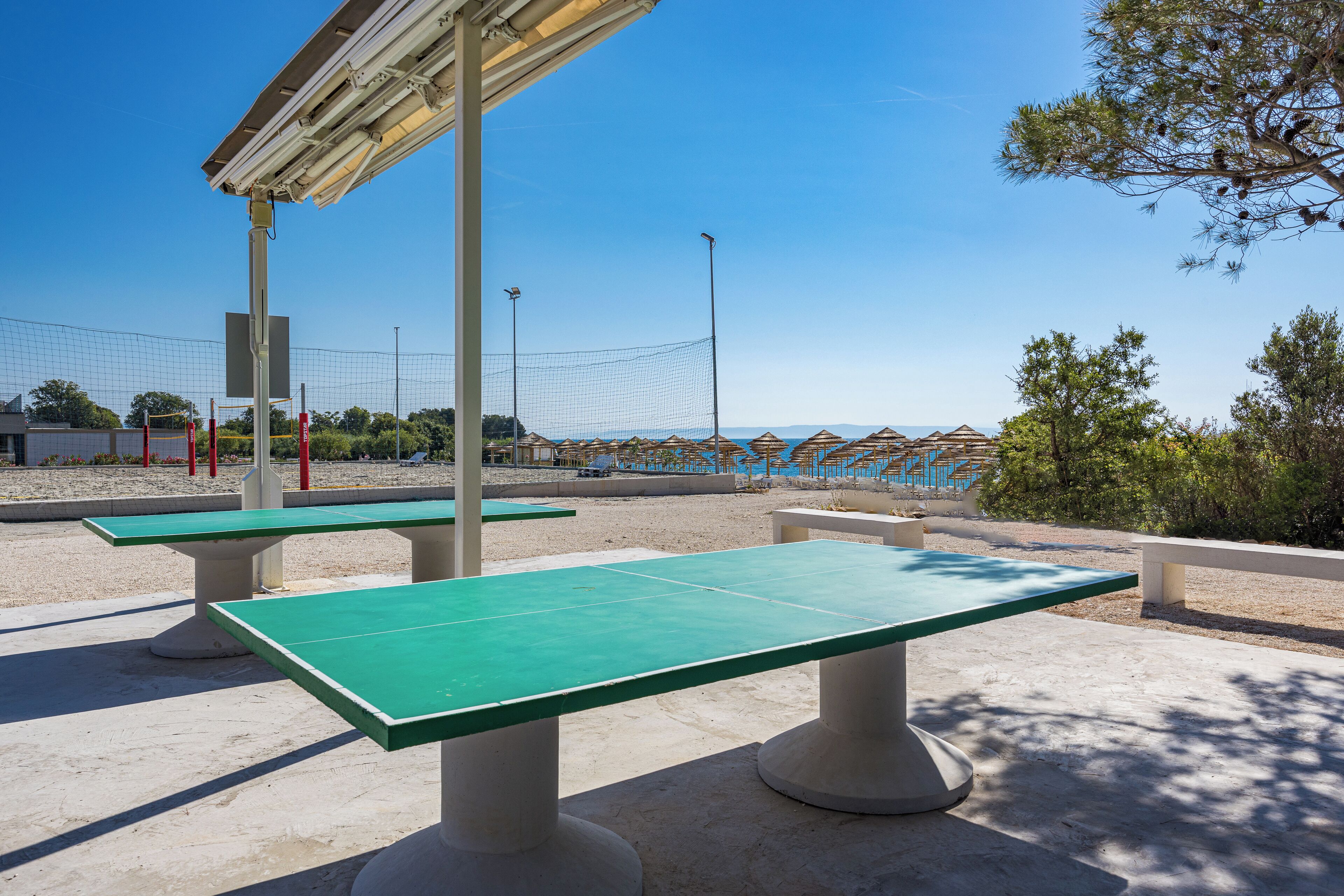 Mesas de ping pong al aire libre bajo una zona cubierta, con vista despejada a la playa y sombrillas de fondo.