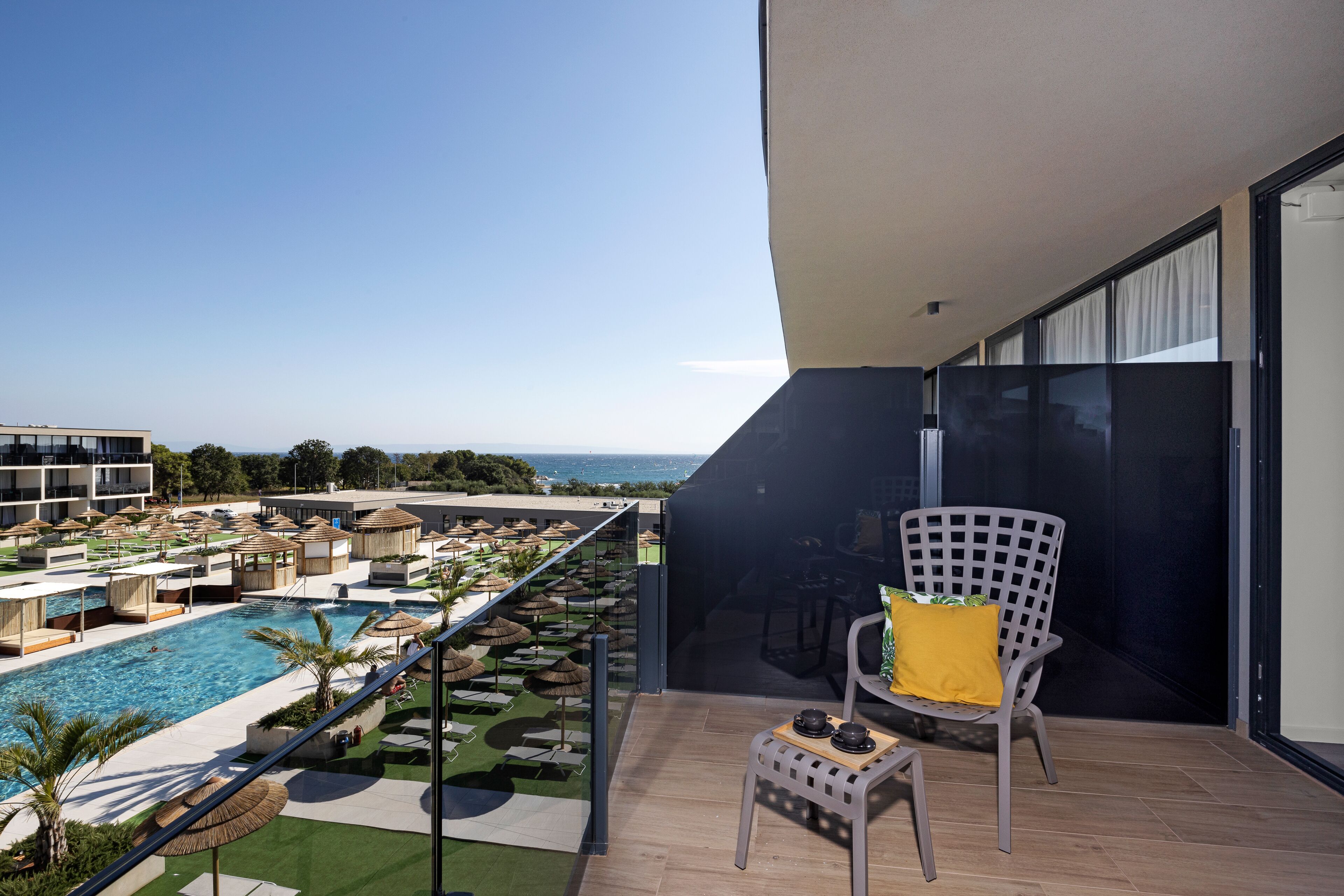 Un balcone con vista su una piscina e un'area relax, una sedia con cuscino giallo e un tavolino con teiera e tazza.