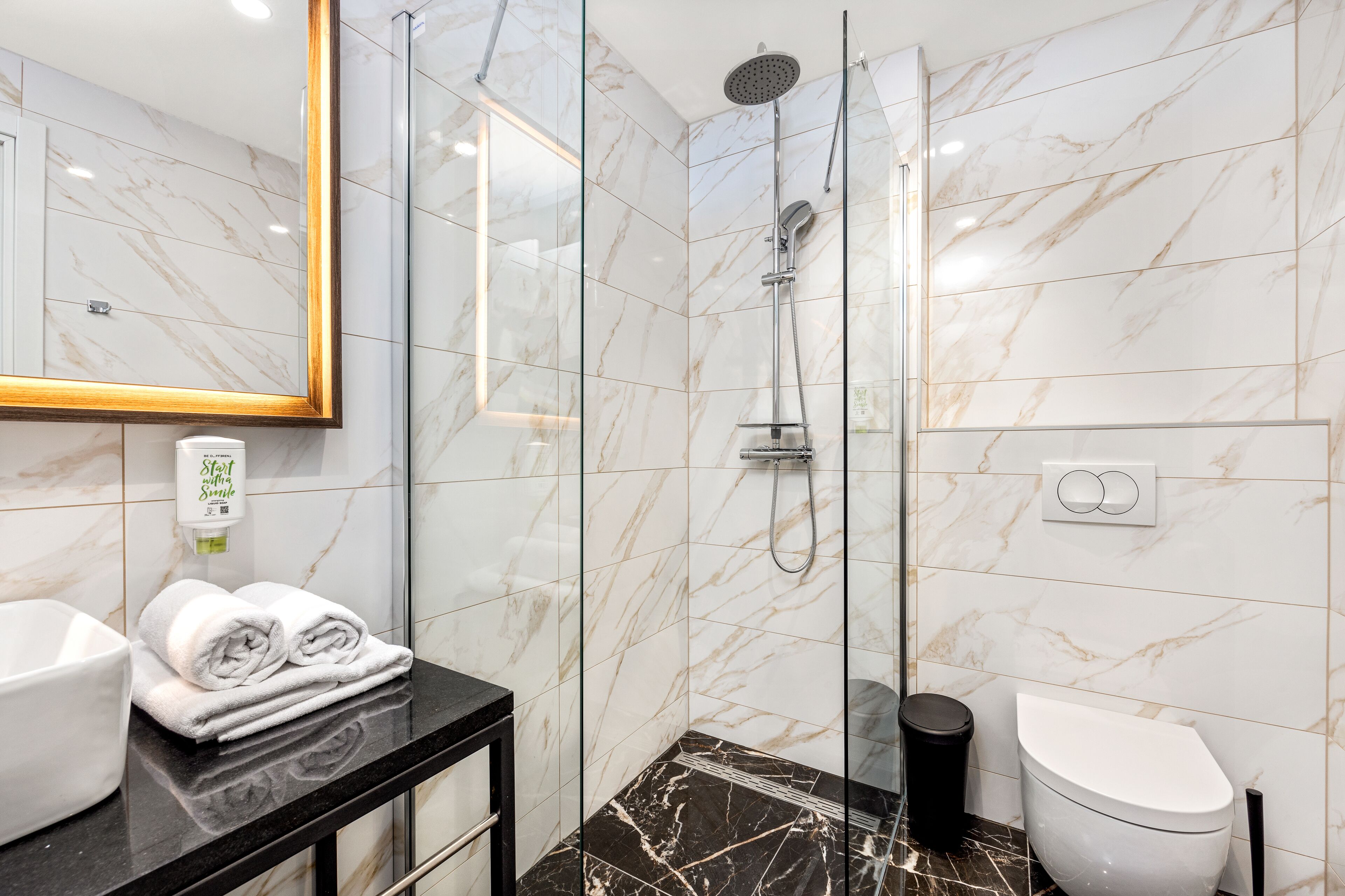 Un bany modern amb dutxa de vidre, rajoles de marbre, un taulell negre, un lavabo blanc i un vàter.