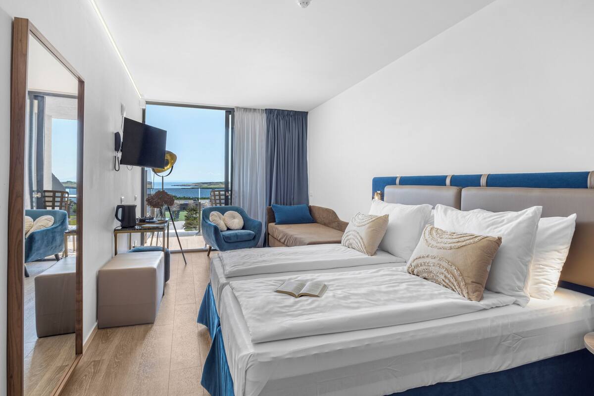 A hotel room with a large bed, a TV, a desk with a lamp, and a view of the sea.