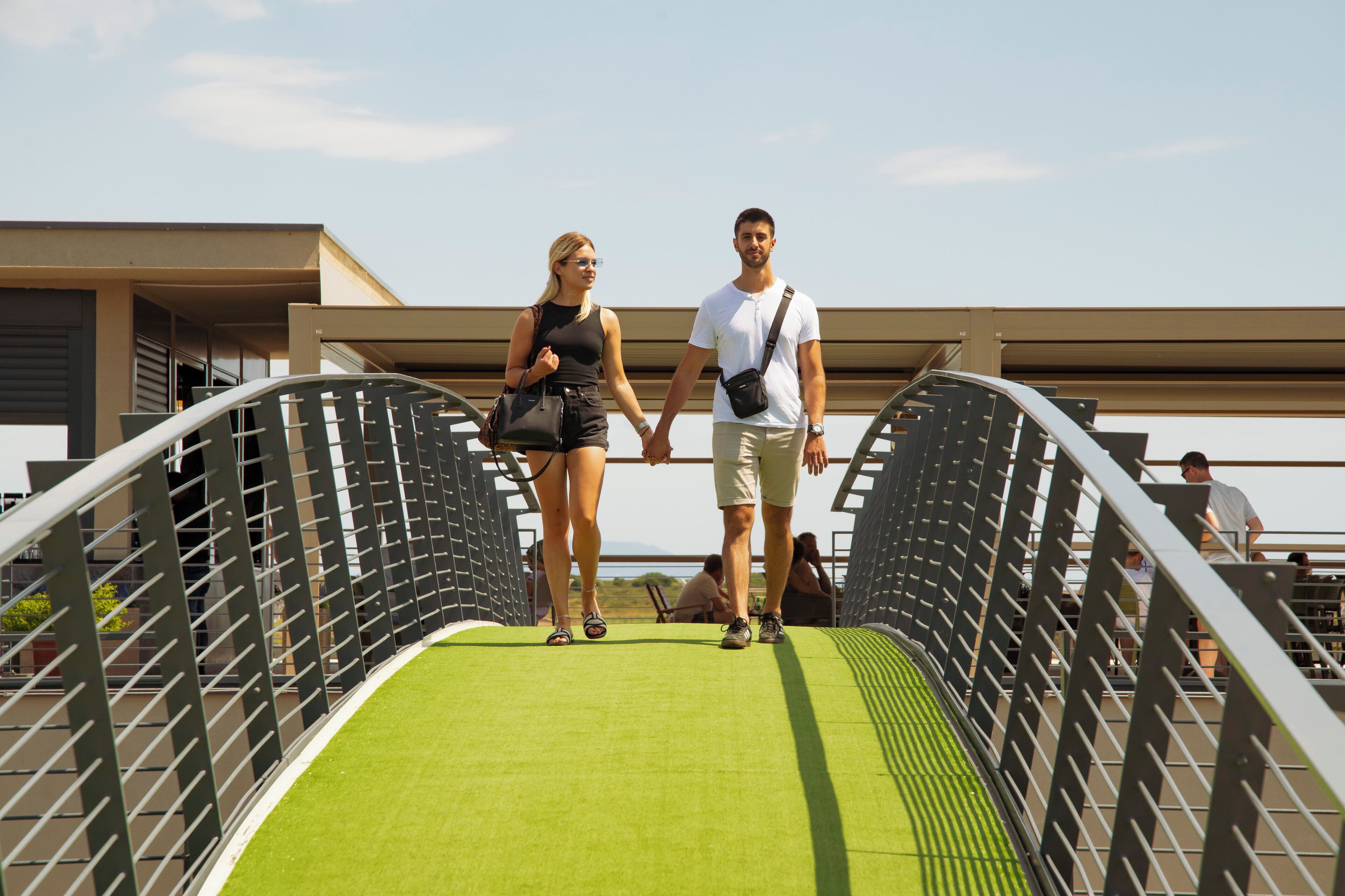 A couple walking hand in hand on a bridge with a green surface, metal railings, and a building in the background.