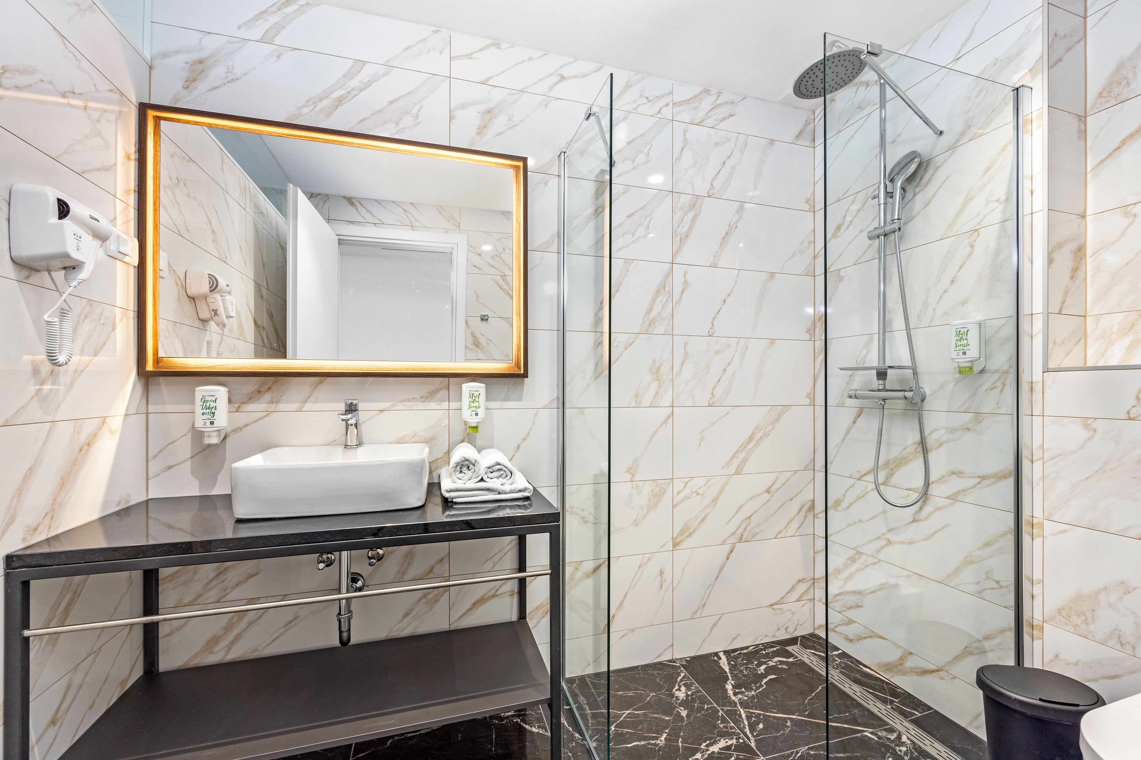 Un bagno moderno con parete rivestita in marmo, un grande specchio con cornice dorata, un lavandino bianco, un mobile bagno nero e un'area doccia con pareti in vetro.
