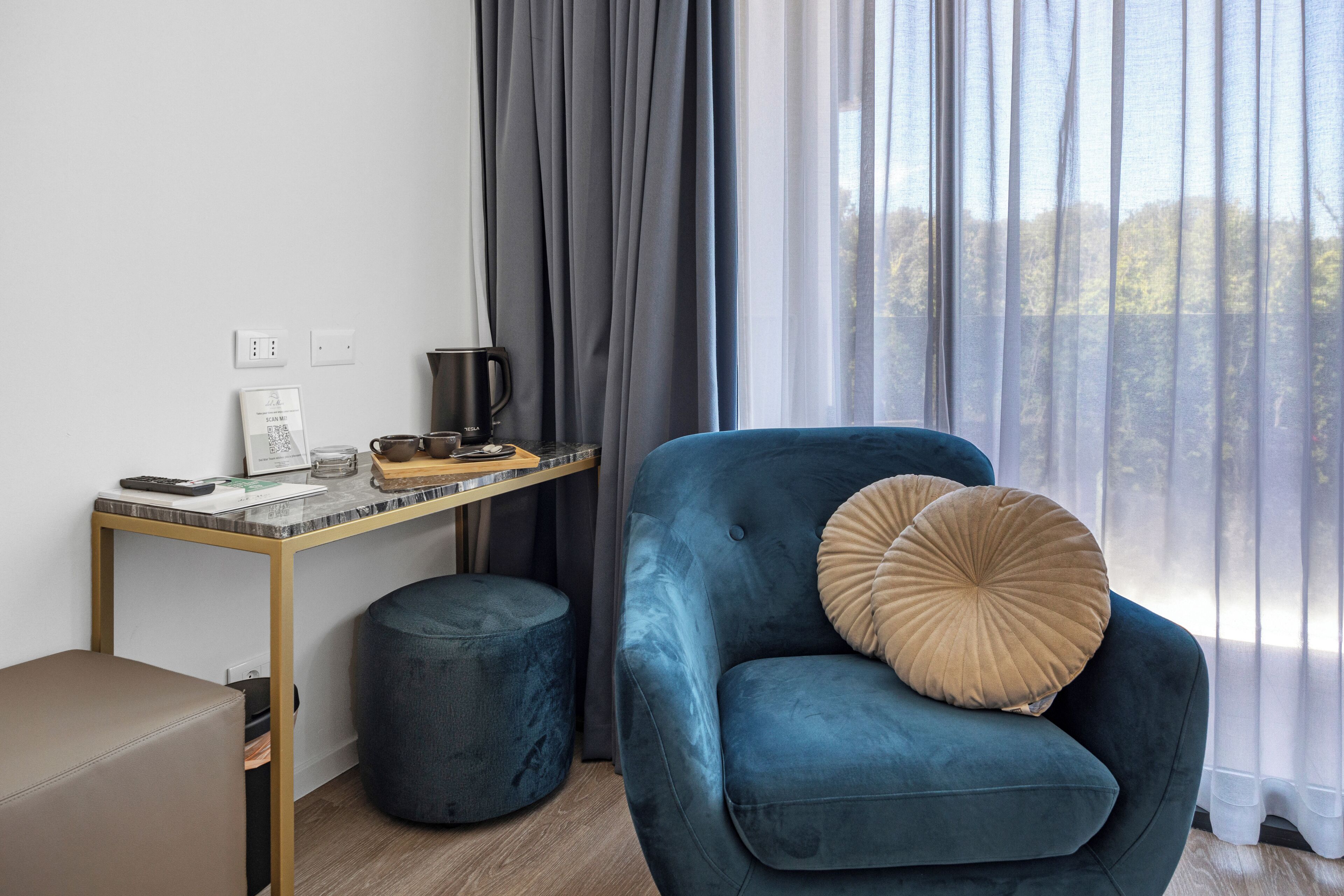 A room with a blue armchair, a small round ottoman, a wooden desk with a coffee maker, and a window with sheer curtains.