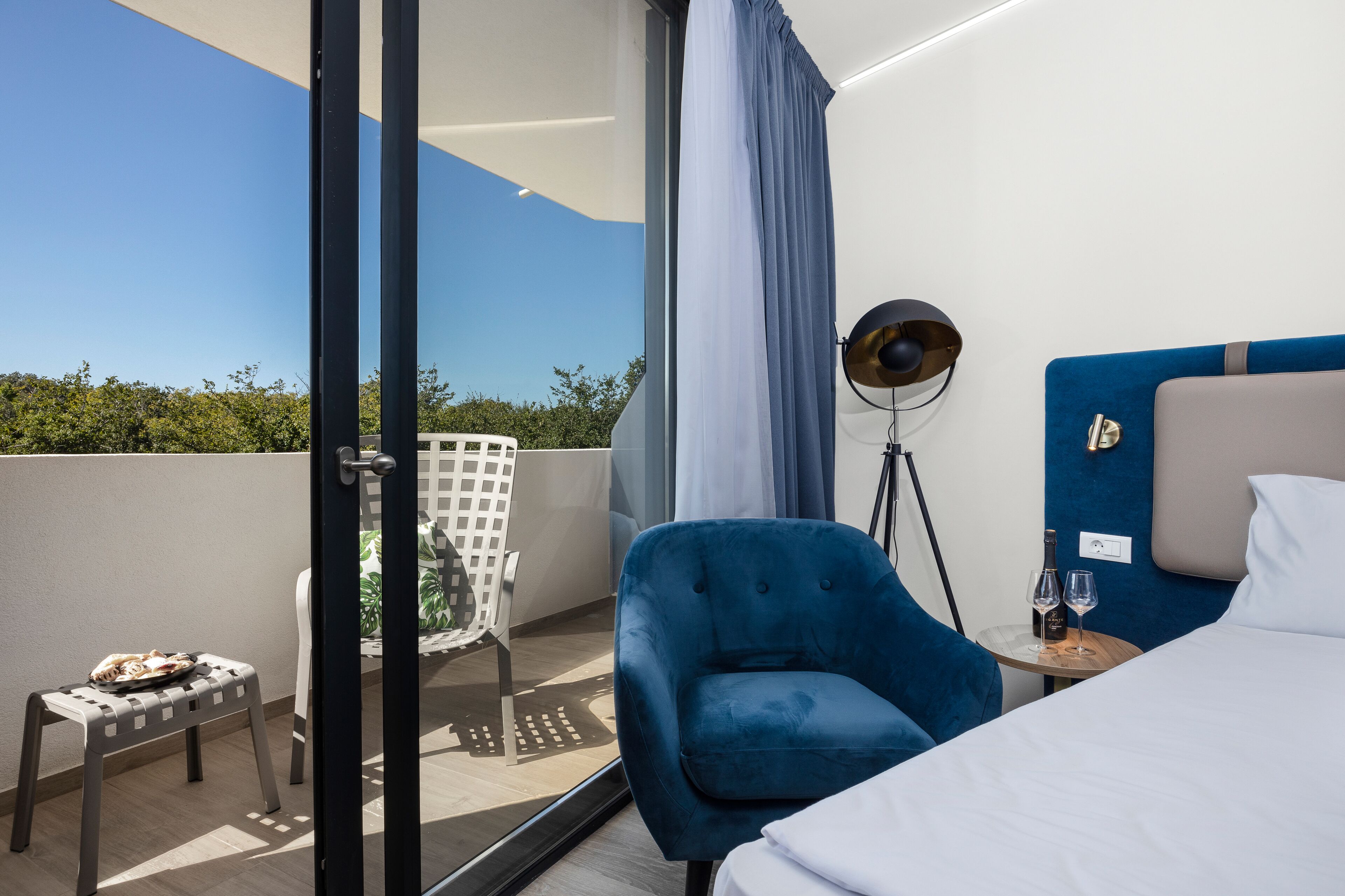 A modern hotel room with a bed, a blue armchair, a small table with a bottle and glass, and a balcony with a chair and a small table.