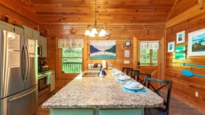 A wooden cabin kitchen with granite countertops, a stainless steel refrigerator, and a dining area with chairs and a table set for four. - A Smoky Mountain Lake Cabin, Spacious Smoky Mountain Getaway, Free Tickets! (Sevierville)