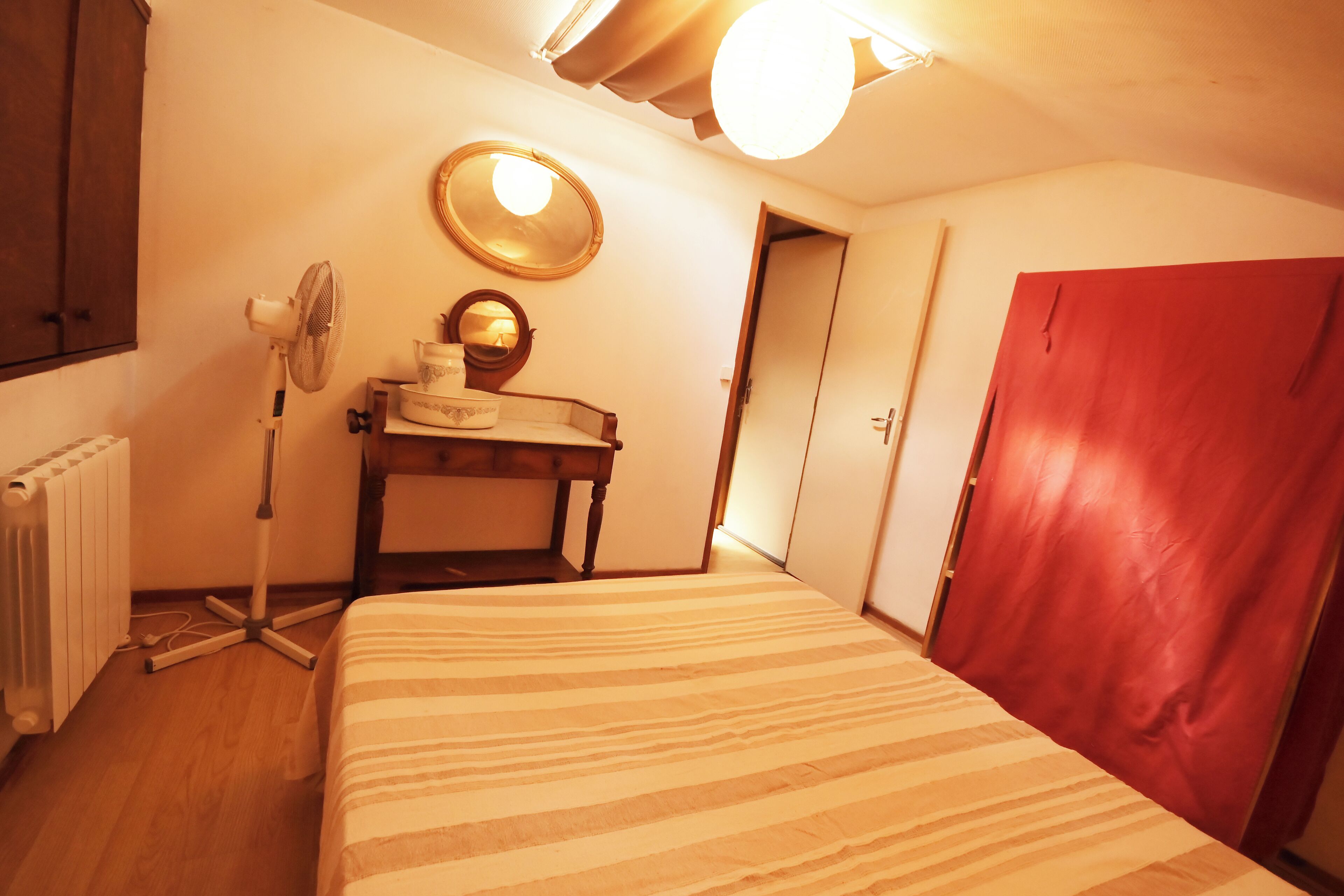 A room with a bed, a desk with a mirror, a radiator, and a red door.