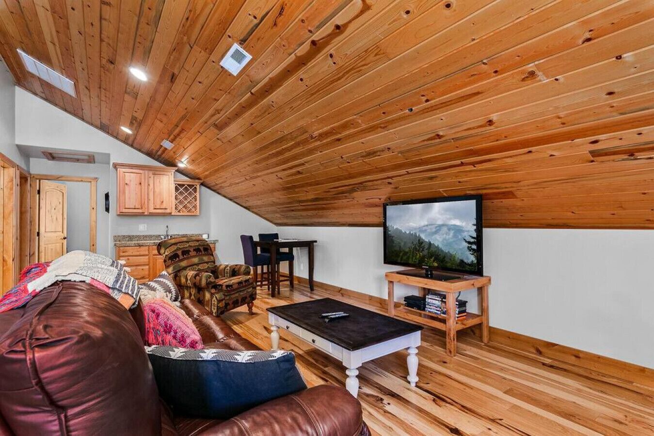 A cozy loft-style room with wooden ceiling beams, a leather sofa, a TV, and a dining area.