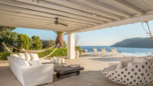 A covered outdoor seating area with white wicker furniture, a hammock, and a view of the sea and hills.