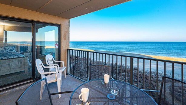 A balcony with a view of the ocean, a dining table with chairs, and a bed with a view.