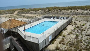 A rectangular swimming pool with a wooden deck and a covered area, surrounded by a sandy beach and a wooden fence.