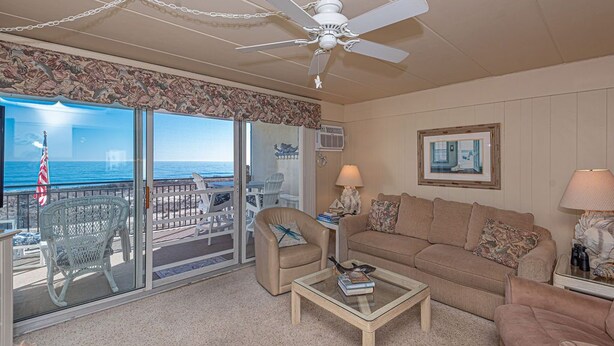 A living room with a beige sofa, a coffee table, and a ceiling fan. There is a view of the ocean through large sliding glass doors.