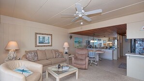 A living room with a beige sofa, a coffee table, a chair, and a ceiling fan. There is a kitchen area visible in the background.