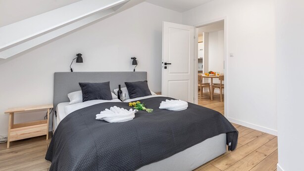 Ein modernes Schlafzimmer mit schräg verlaufender Decke, einem großen Bett mit dunkler Bettdecke, zwei Nachttischlampen und einem kleinen Holfnachtkästchen.