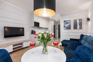 A modern living room with a flat-screen TV, a round coffee table with two wine glasses, a vase with flowers, and a blue sofa. - Stylish Studio with Balcony | Jacuzzi, Parking (Dziwnów)