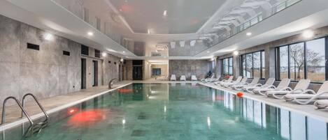 A large indoor swimming pool with a clear water surface, surrounded by lounge chairs and a modern architectural design.