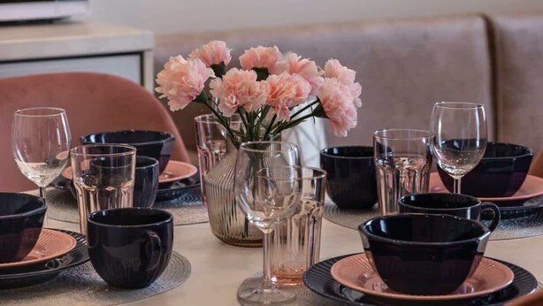Ein Esstisch mit schwarzen und rosafarbenen Tellern, klaren Gläsern und einer Vase mit rosafarbenen Blumen.