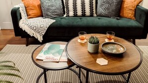 A cozy living room with a dark green sofa adorned with patterned cushions, a small round coffee table with a plant and a bowl, and a magazine on a patterned rug.