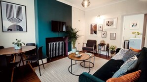 A living room with a sofa, coffee table, and chairs. There is a dining table with chairs, a radiator, and a television on the wall.