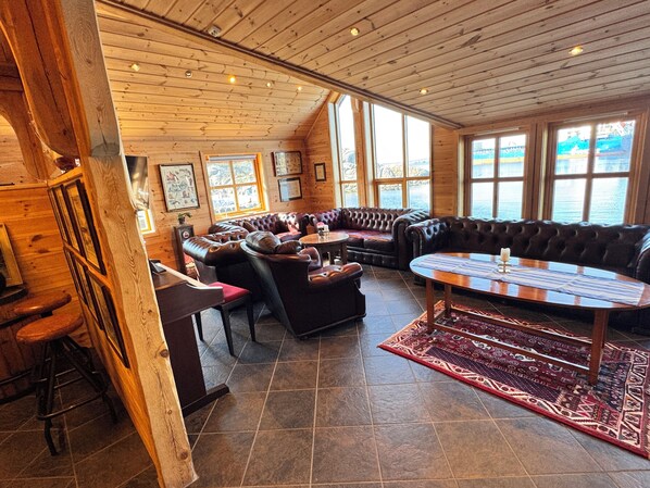 A wooden interior with a large leather sofa, a round table, and a view through large windows. - Sirevåg Hotel (Ha)