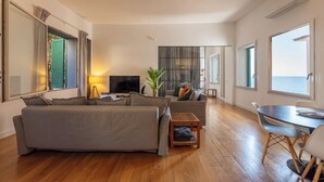 Ein modernes Wohnzimmer mit einer Couch, einem Fernseher, einem kleinen Tisch und Blick aufs Meer.