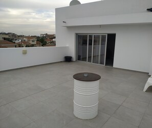 Una terraza en la azotea con un objeto cilíndrico blanco, una puerta corrediza de vidrio y vistas de edificios circundantes.
