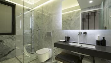 A modern bathroom with a glass-enclosed shower, a white toilet, a large mirror, and a dark countertop with toiletries.