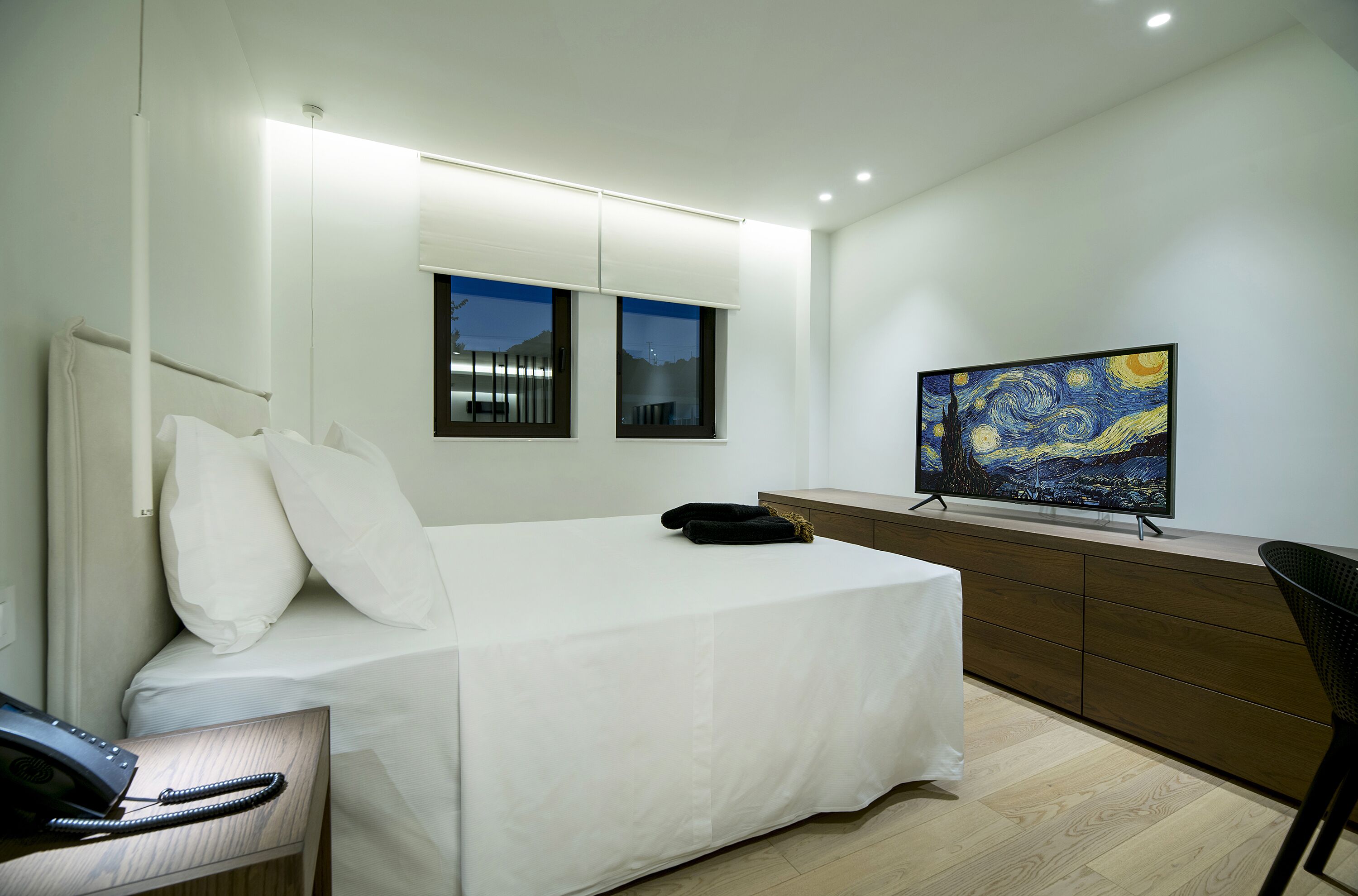 Ein modernes Hotelzimmer mit einem ordentlich bezogenen Bett, einem Flachbildfernseher, einem Schreibtisch mit Telefon und einem großen Fenster mit Nachtblick.