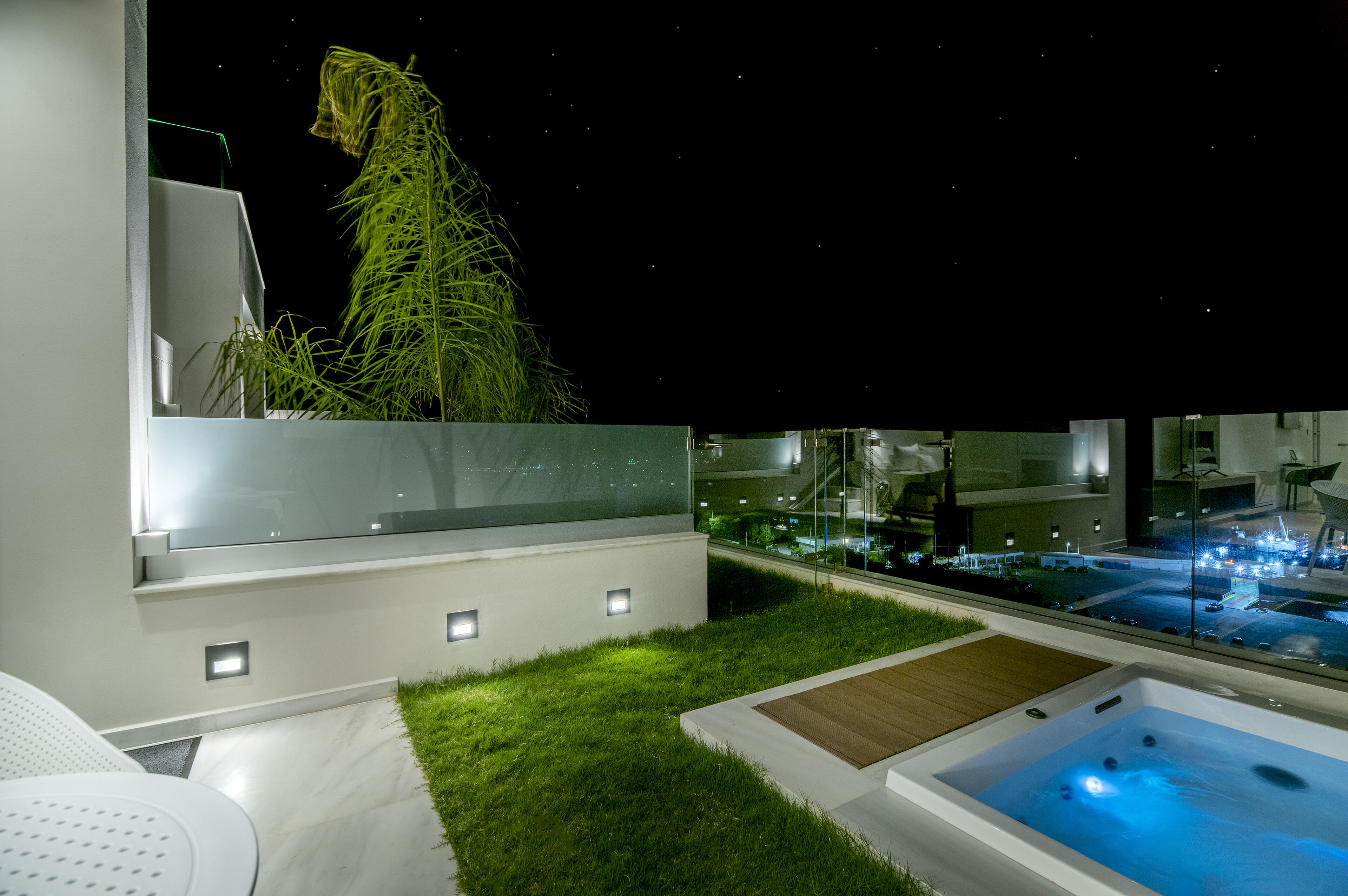 Un espacio exterior moderno con barandilla de cristal, una bañera de hidromasaje y un césped cuidado.