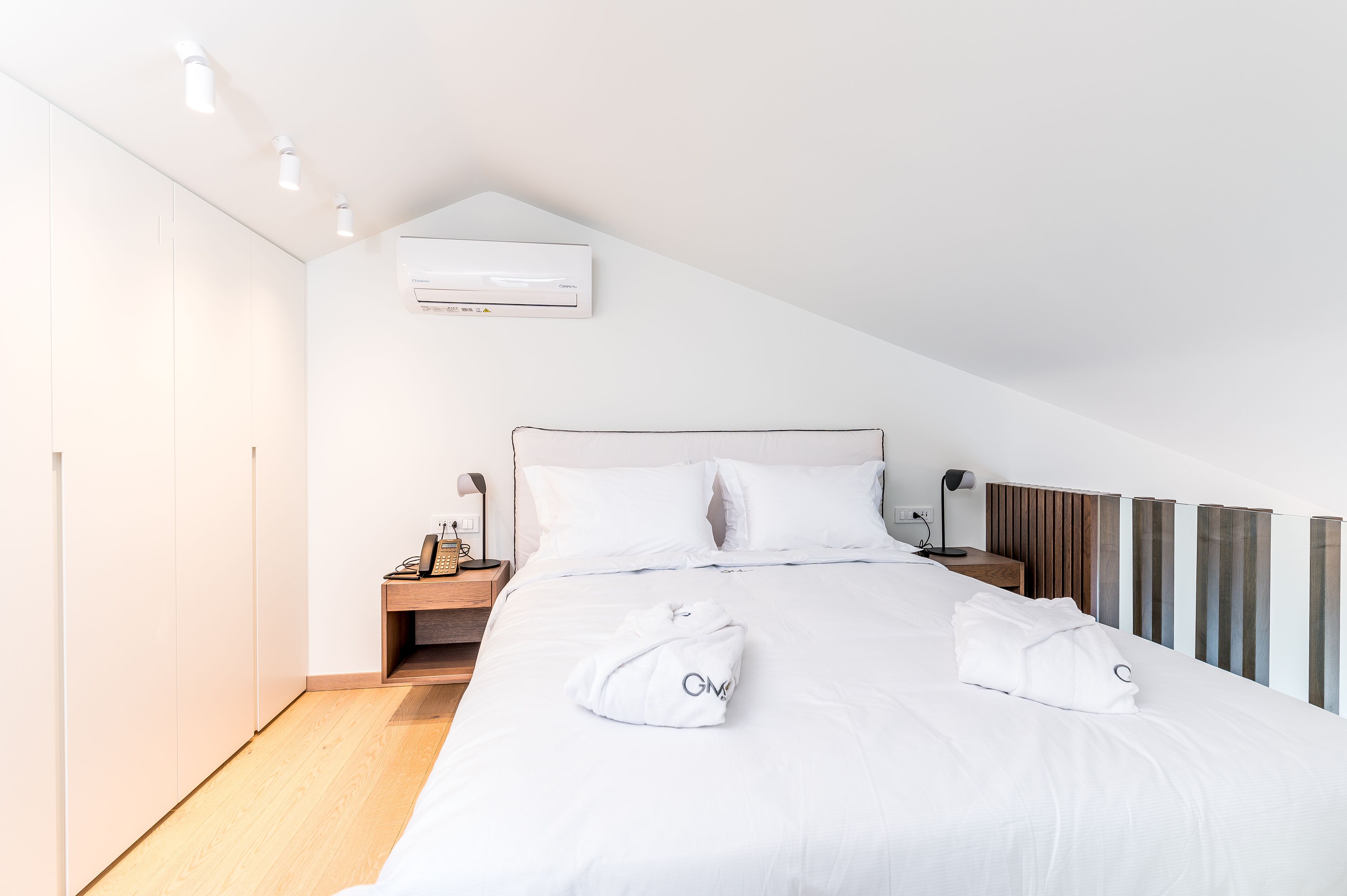 Une chambre d’hôtel moderne avec un grand lit, du linge de lit blanc, une table de chevet en bois, une lampe et un climatiseur fixé au mur.