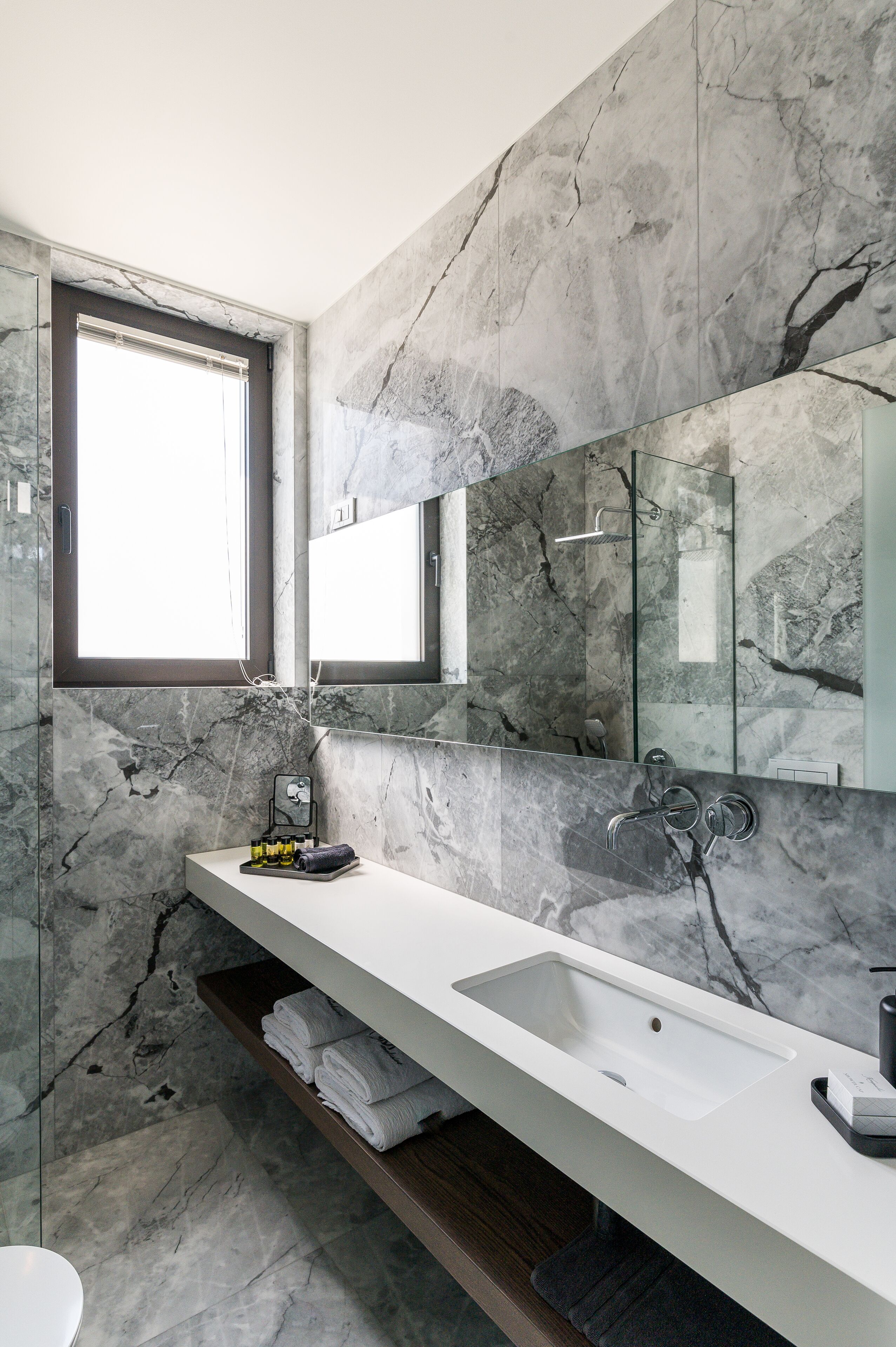 A modern bathroom with a marble wall, a large mirror, a white sink, and a towel rack.