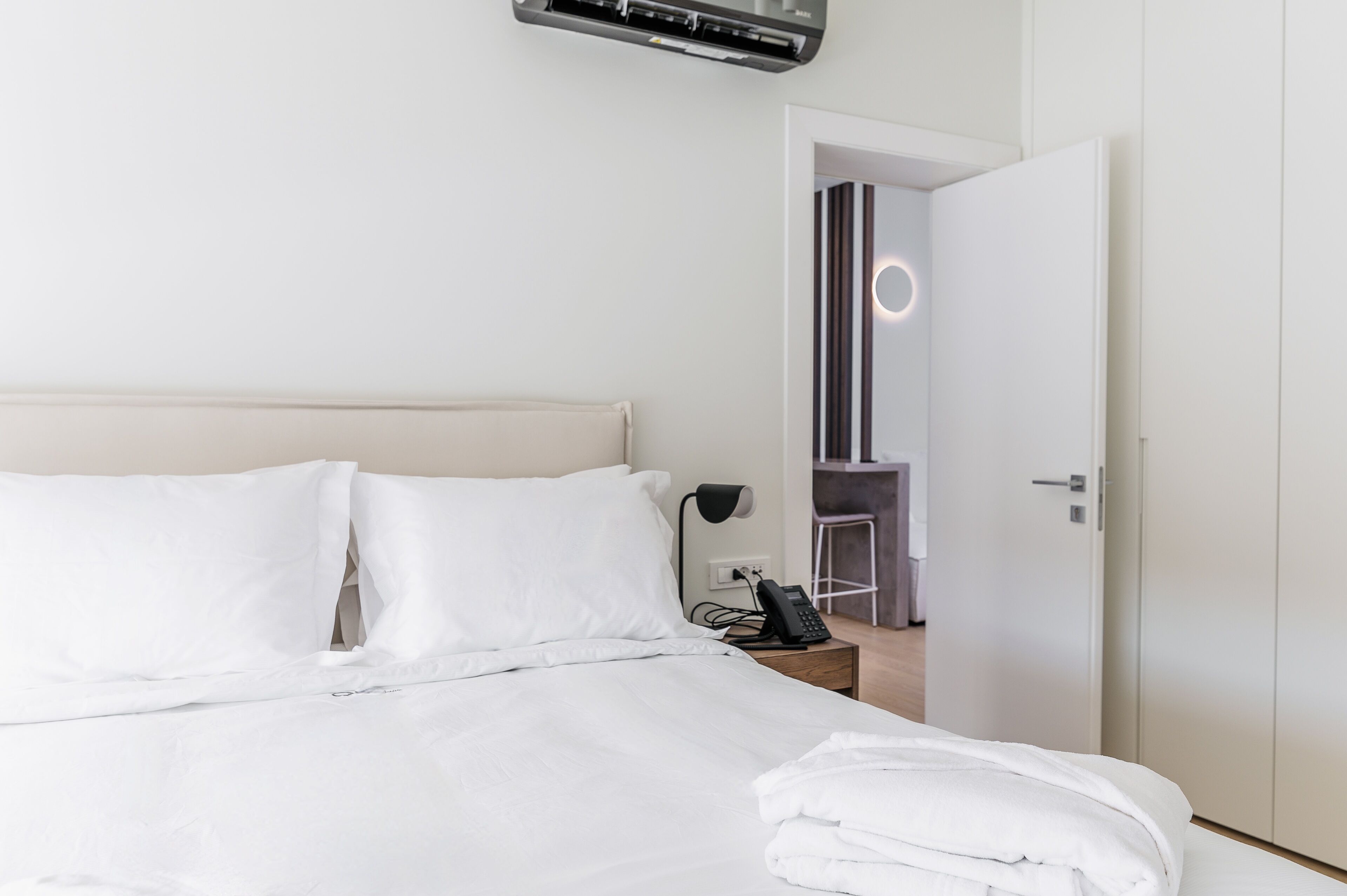 A neatly made bed with white linens, a bedside phone, and a wall-mounted air conditioner.