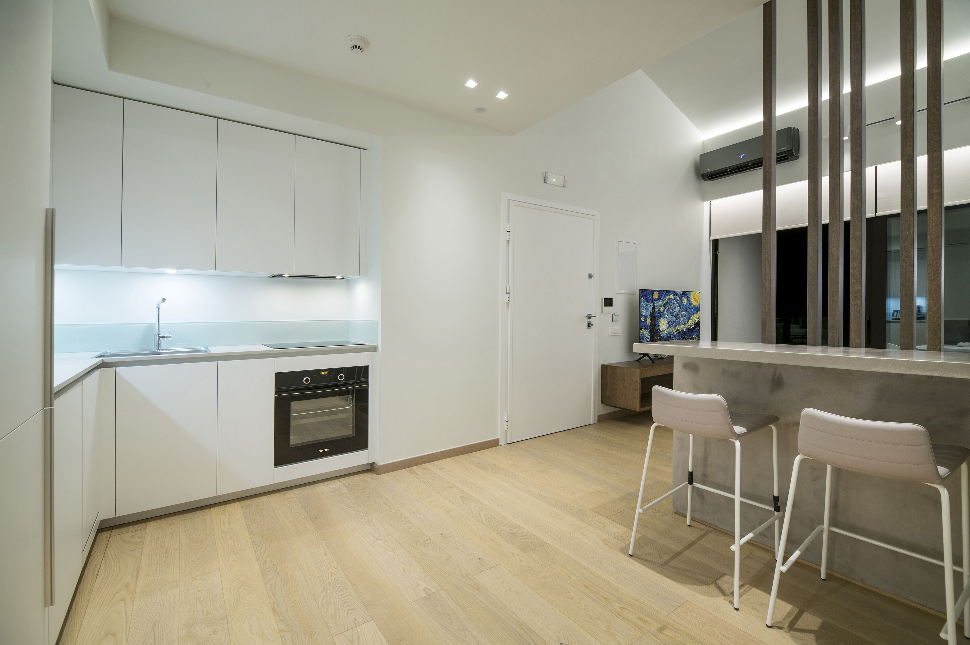 A modern kitchen with white cabinets, a built-in oven, and a bar area with two white bar stools.
